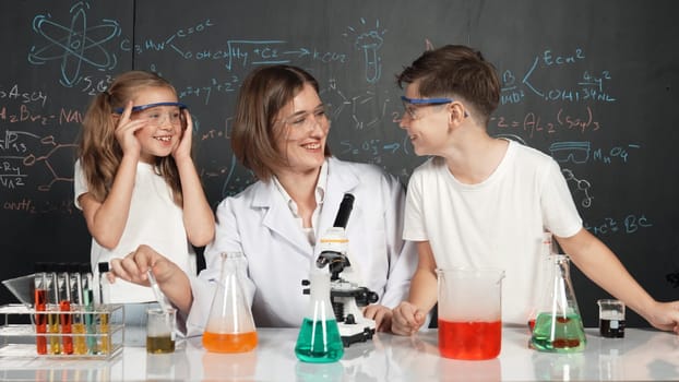 Caucasian boy mixing chemical liquid while teacher giving advice. Professional instructor wearing lab suit looking for diverse student at table with beaker filled with colored solution. Erudition.