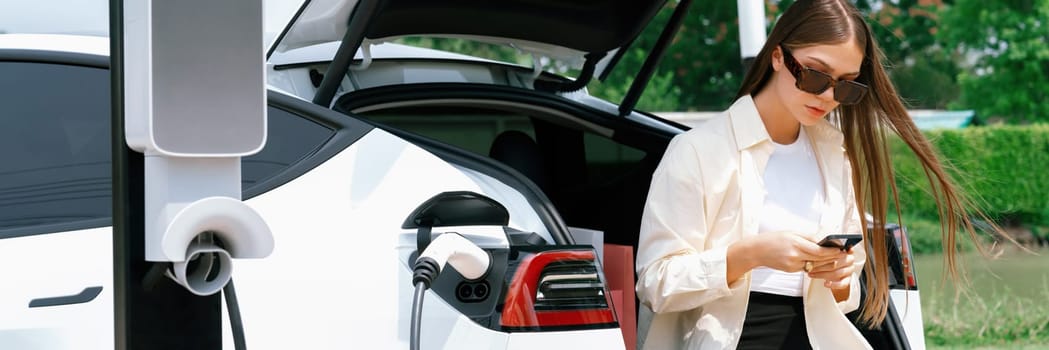 Young woman use smartphone to pay for electricity at public EV car charging station green city park. Modern environmental and sustainable urban lifestyle with EV vehicle. Panorama Expedient