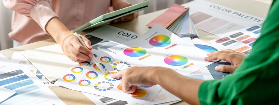 Skilled architect searching data from tablet while selecting an appropriate color from color wheel at table with color palette and document scatter around. Creative design concept. Variegated.