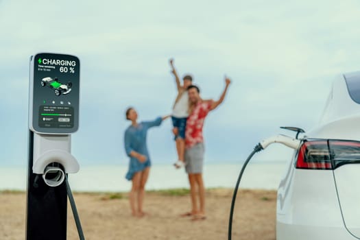 Alternative family vacation trip traveling by the beach with electric car recharging battery from EV charging station with blurred cheerful and happy family enjoying the seascape background. Perpetual