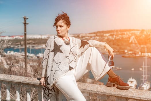 Woman walks around the city, lifestyle. A young beautiful woman in white trousers and a sweater sits on a white fence with balusters and overlooks the sea bay and the city