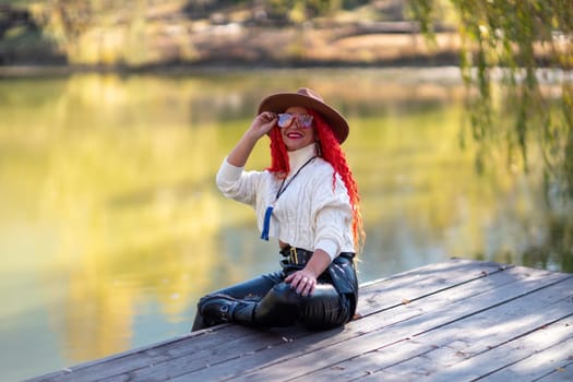 Autumn lake woman. In autumn, she sits by the pond on a wooden pier and admires nature with red hair and a hat. Tourism concept, weekend outside the city