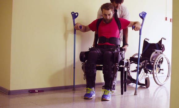 Young disable man in the robotic exoskeleton sitting in wheelchair in the rehabilitation clinic