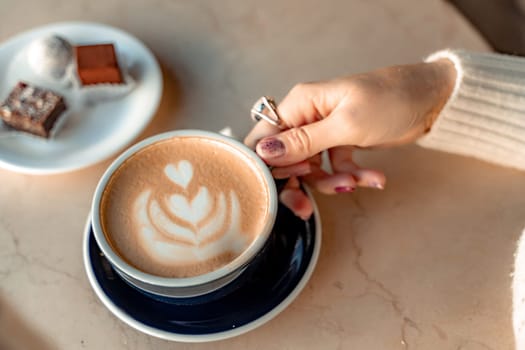 A cup of fresh cappuccino coffee in the hands of a woman on a fashionable background of a white marble table, next to a plate with sweets. Coffee addiction. Top view, flat lay