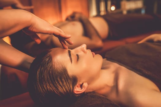 Couple customer enjoying relaxing anti-stress head massage and pampering facial beauty skin recreation leisure in warm candle lighting ambient salon spa in luxury resort or hotel. Quiescent