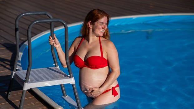 A red-haired pregnant woman goes down the stairs into the pool outdoors