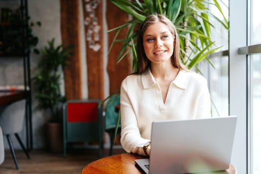 A young woman with a pleasant smile is seated at a wooden table in a well-lit, contemporary cafe. Shes engaged in her work on a laptop, dressed in casual professional attire, and appears to be confidently managing her tasks.