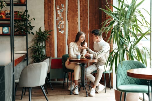 A smiling young couple sits closely together at a small table in a warmly lit cafe, sharing a moment over coffee. Surrounded by indoor plants and rustic decor, they seem engaged in a pleasant conversation, with a backdrop of draping curtains and a casual atmosphere.