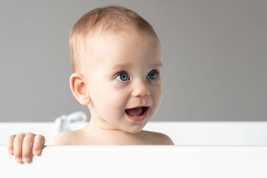 Close-up of the face of a baby laughing out loud and looking out of a crib with interest.
