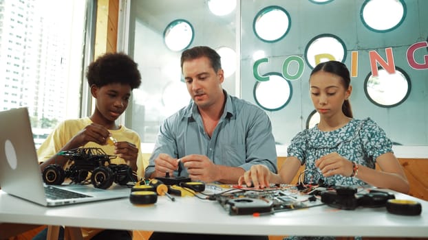 Skilled teacher teaching diverse students fixing car model by using wire and electronic equipment. Girl studying about using electrical tool to check mother board while looking at laptop. Edification