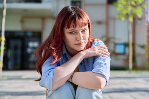 Upset sad unhappy young female sitting on steps. Pensive serious red-haired girl university college student looking at camera, outdoor. Problems difficulties depression mental health of young people