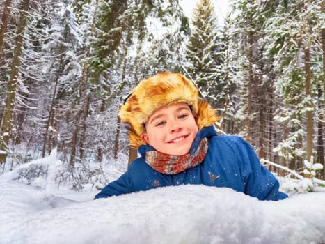 Funny boy in the snow in a snowdrift in a Russian hat with earflaps. A child is playing at war. A future soldier. The war between Russia and Ukraine