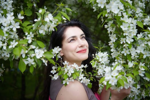 Joyous brunette woman near Blossoms of apple tree in a Spring Garden outdoors. The Concept of face and body care. The scent of perfume and tenderness