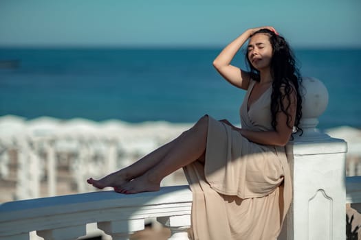 A woman in a tan dress is sitting on a white railing overlooking the ocean. The scene is serene and peaceful, with the woman enjoying the view and the calming sound of the waves