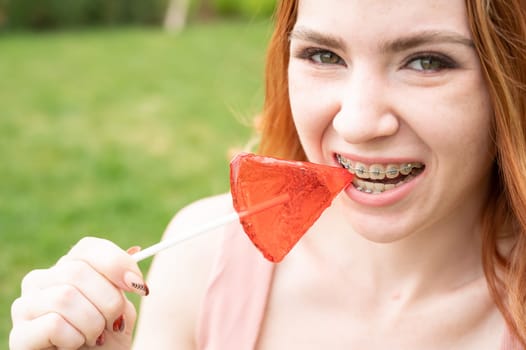 Beautiful young woman with braces on her teeth eats a watermelon-shaped lollipop outdoors