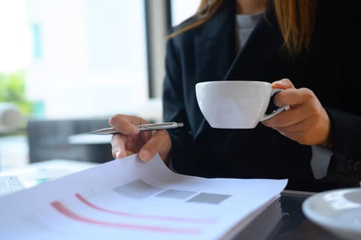 Professional businesswoman holding cup of hot coffee and checking financial reports at desk.