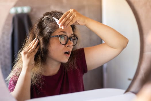 Woman Looking for Gray Hair in the Mirror. Young woman scared of premature aging signs.