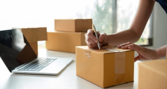 A small business owner receives product orders and writes shipping information on cardboard boxes in the home office to prepare for delivery..