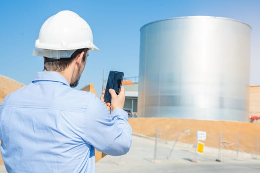 An engineer wearing a hard hat is using his cell phone to take a picture of a tank at a gas or water facility. He is dressed in workwear and a composite material helmet