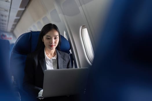 A woman is sitting in an airplane seat with a laptop open in front of her. She is wearing a suit and she is working on her laptop. Concept of productivity and focus