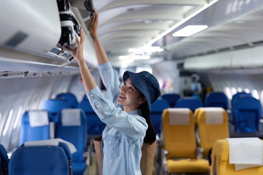 A woman is reaching for something in the overhead compartment of an airplane. She is wearing a blue shirt and a blue hat