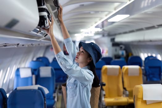 A woman is reaching for something in the overhead compartment of an airplane. She is wearing a blue shirt and a blue hat