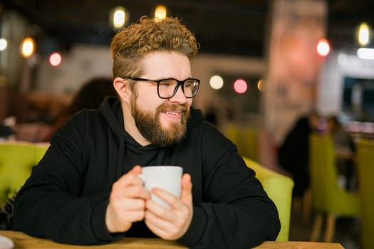 Bearded curly man smiling confident drinking coffee in restaurant or coffee shop. Millennial generation and Gen Y.
