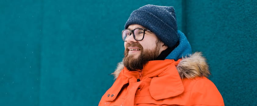 Portrait handsome eyeglasses young male smiling bearded stands on a blue wall background in a bright orange winter jacket with a hood with fur in winter. Millennial generation and Gen Y