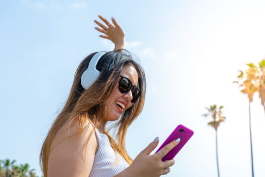 Excited Chinese girl in trendy sunglasses in headphones enjoying music outdoor on summer resort. High quality photo