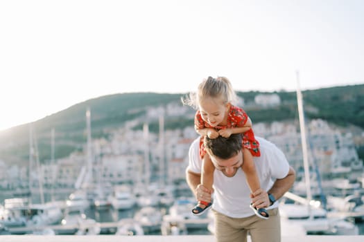 Dad with a smiling little girl on his shoulders stands leaning forward on the seashore. High quality photo