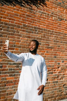 Happy young African American man in dashiki ethnic clothes taking selfie on brick wall background. Millennial generation student and youth