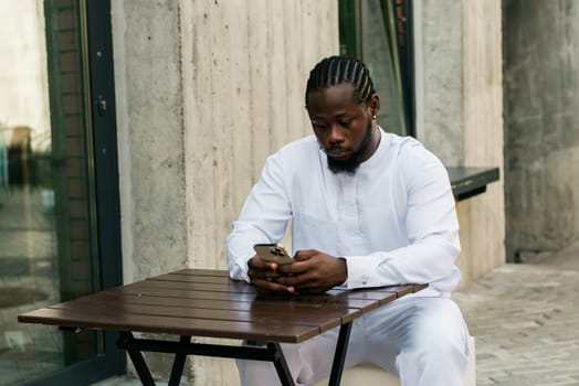 African American hipster guy in cultural and ethnic clothing dashiki chatting on smartphone outdoors, texting with friends on social networks. Millennial and Internet concept.