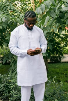 African American hipster guy in cultural and ethnic clothing dashiki chatting on smartphone outdoors, texting with friends on social networks. Millennial and Internet concept.