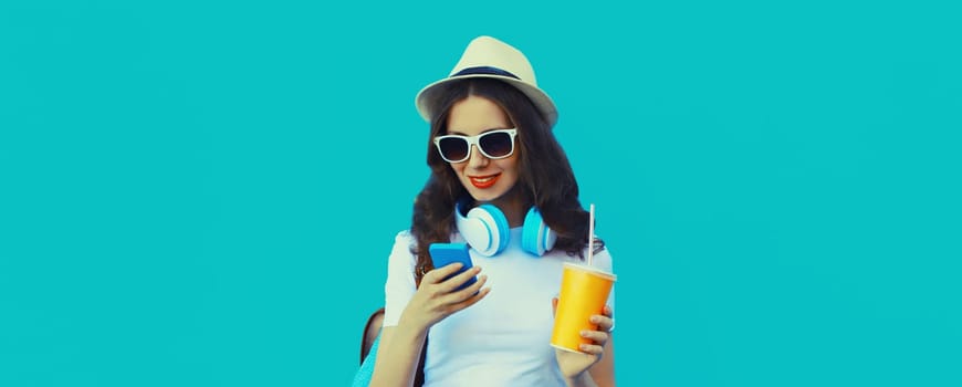 Portrait of modern young woman listening to music with headphones holding phone and cup of fresh juice on blue studio background