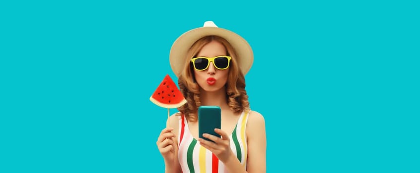 Summer portrait of happy young woman holding smartphone with juicy lollipop or ice cream shaped slice of watermelon wearing straw hat on blue background