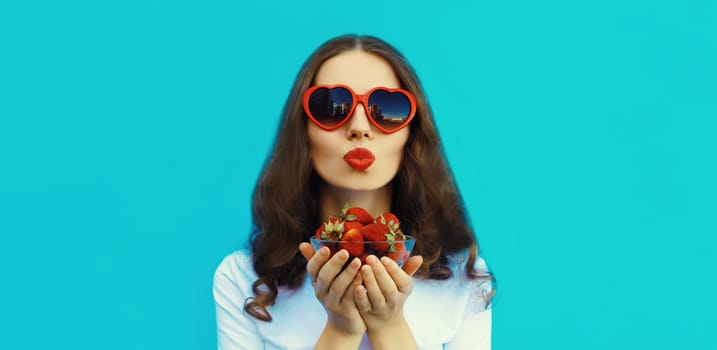 Portrait of beautiful caucasian young woman with handful of fresh strawberries blowing kiss wearing red heart shaped sunglasses on blue studio background