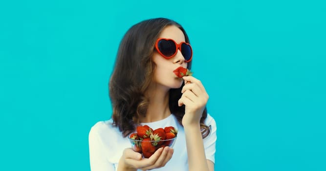 Portrait of beautiful caucasian young woman with handful of fresh strawberries blowing kiss wearing red heart shaped sunglasses on blue studio background