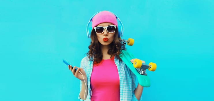Portrait of stylish modern happy young woman listening to music in headphones with mobile phone and skateboard posing wearing colorful clothes, pink hat, sunglasses on blue studio background