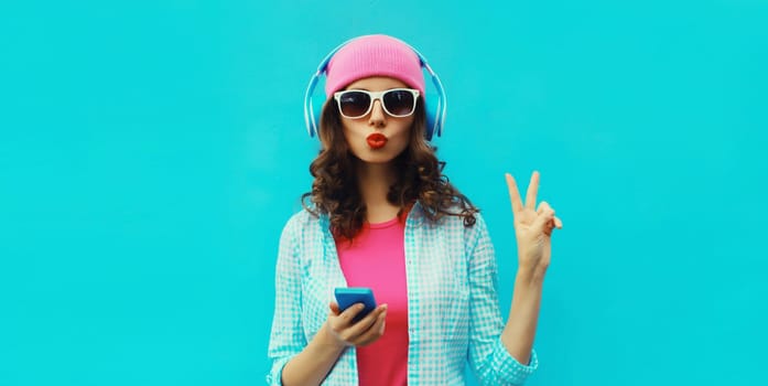 Portrait of stylish modern happy young woman listening to music in headphones with mobile phone wearing pink hat, sunglasses on blue studio background