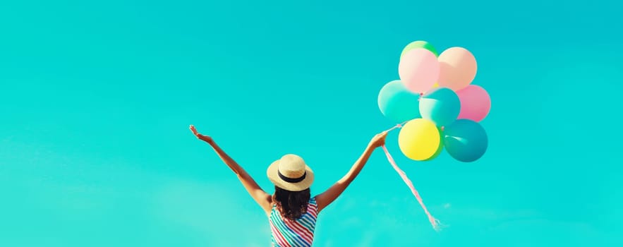 Back view of happy joyful young woman with bunch of colorful balloons in summer straw hat on blue sky clouds background