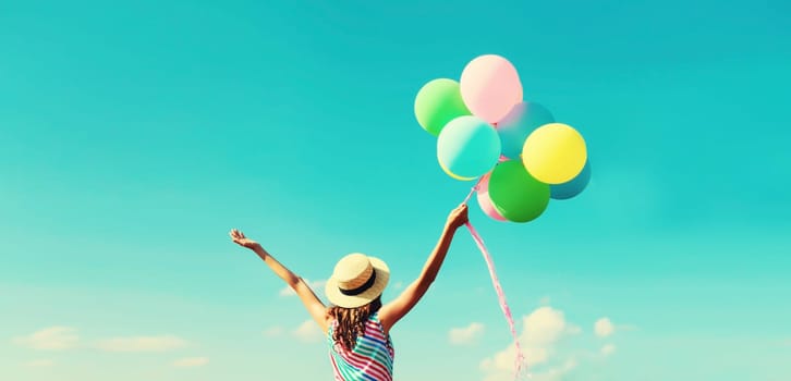 Back view of happy joyful young woman with bunch of colorful balloons in summer straw hat on blue sky clouds background