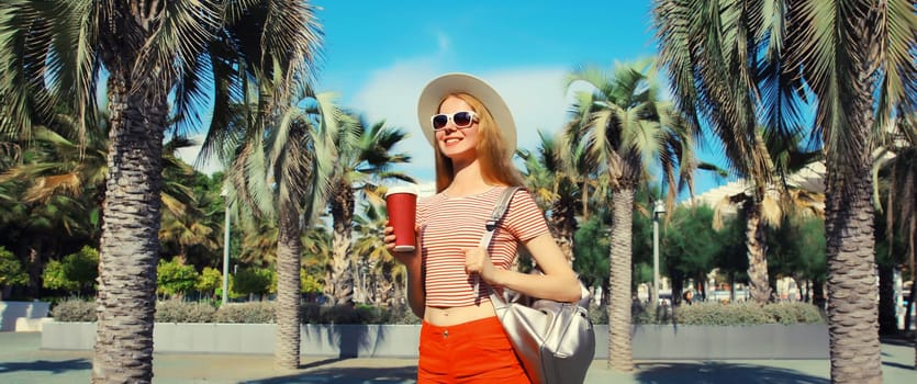 Beautiful young woman with cup of coffee in summer park wearing backpack, straw hat, shorts against a palm trees background