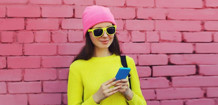 Modern caucasian young woman 20s model with mobile phone looking at device wearing colorful vivid clothes, yellow sweater and pink hat on city street