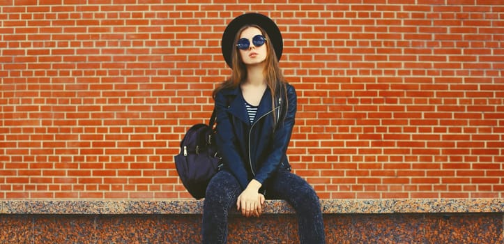 Portrait of stylish young woman model wearing black round hat, leather jacket in rock style on city street on brick wall background