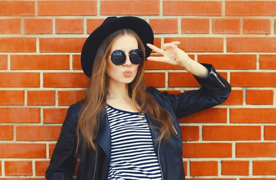 Portrait of stylish young woman model wearing black round hat, leather jacket in rock style on city street on brick wall background