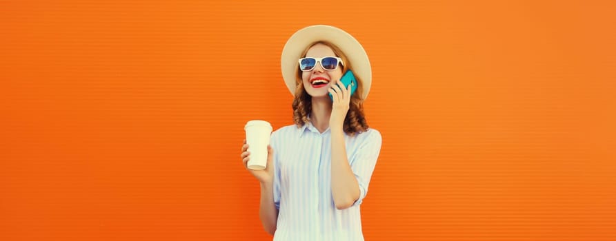 Happy cheerful smiling young woman calling on mobile phone with cup of coffee wearing summer straw hat on orange background