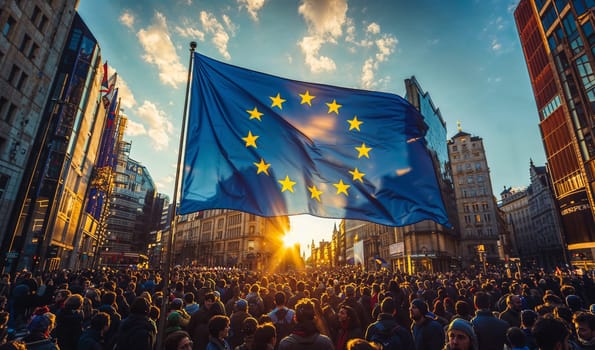 A large gathering of people fills a city square, a European Union flag waving prominently as the sun sets in the background