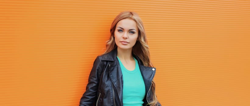 Portrait of beautiful young blonde woman posing in black leather jacket looking at camera on colorful orange background