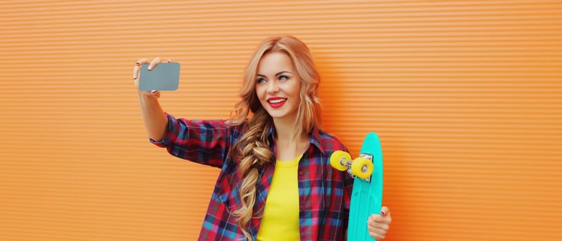 Summer portrait of happy smiling blonde young woman taking selfie with smartphone and skateboard on colorful orange background