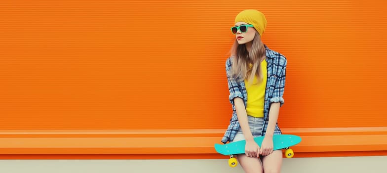 Summer portrait of stylish young woman with skateboard in colorful clothes, yellow hat looking away posing on orange background, blank copy space